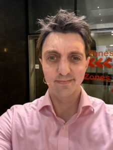 Matt Fricker, an ambassador for Stagetext, is a white male with dark brown hair wearing a pink shirt and a microphone, smiling at the camera