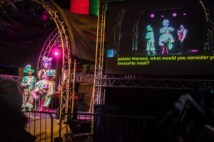 Image of a stage to the left and a screen with live subtitles to the right. The people pon staget are two panto dames and a man